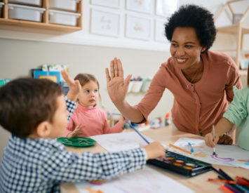 Happy African American teacher and small boy giving high-five during art class at kindergarten.